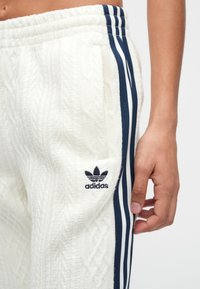 Κοντινό πλάνο ενός χεριού δίπλα σε λευκό πλεκτό παντελόνι Adidas με τρεις ρίγες σε ναυτικό μπλε και λογότυπο στην πλαϊνή τσέπη.