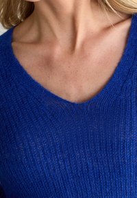 Sweter z dekoltem w serek w intensywnym niebieskim kolorze, z teksturowanym wzorem dzianiny. Tkanina wydaje się lekka i lekko przezroczysta.