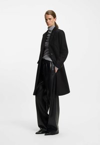 Sort uldfrakke over en grå turtle neck, parret med skinnende sorte plisserede bredde bukser og spidse sorte sko. Modellen står selvsikkert.
