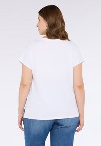 Camiseta blanca de algodón con mangas cortas, corte relajado y escote redondeado, combinada con jeans de denim azul. La vista trasera muestra una textura suave.