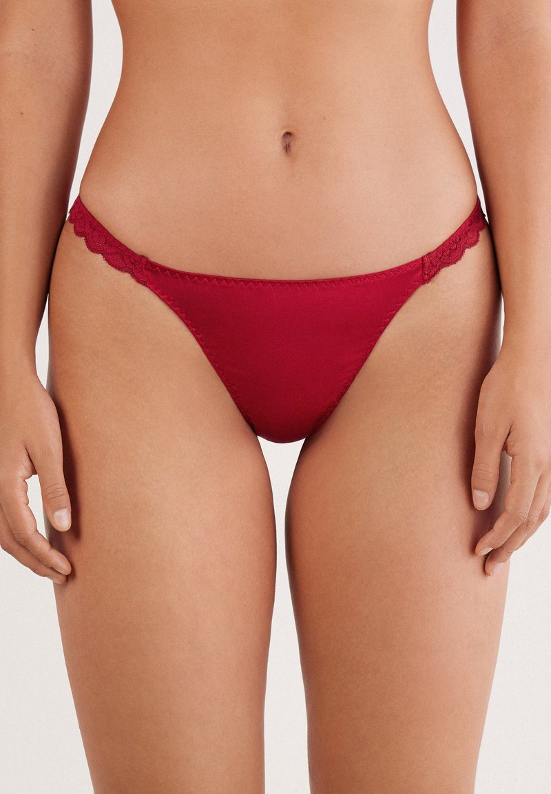 Rød lavtaljet bikininnederdel med blonder, båret af en kvinde, der viser hendes mellemgulv og hænder ved siden af på en ensfarvet baggrund.