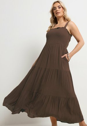 Kobieta w zwiewnej, bezrękawnej, brązowej sukni maxi na piętra z falbanką i ręką w kieszeni, pozująca na jednolitym tle.