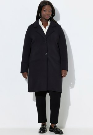 Zwarte jas met reverskraag en knoopsluiting, lange mouwen en een rechte pasvorm, gecombineerd met zwarte broek en loafers.