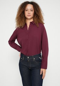 Bordová košeľa s dlhým rukávom a zapínaním na gombíky vpredu, kombinovaná s tmavými džínsami klasického strihu s výraznými švami.