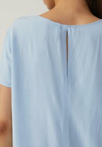 Haut à manches courtes bleu clair avec un col rond, une fente dans le dos, et un tissu lisse avec un léger éclat.