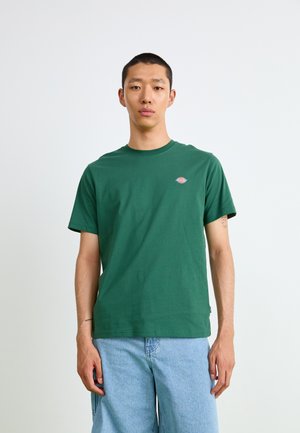 Green cotton T-shirt with a round neckline, short sleeves, and a small logo on the left chest; paired with light blue jeans.