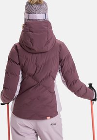 Violette und weiße gepolsterte Skijacke mit Kapuze, diagonal gesteppt und mit einem orangefarbenen Logoetikett. Ein Skistock wird in der rechten Hand gehalten.