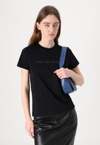 Juodas medvilnės marškinėliai su toniniais „KARL LAGERFELD“ užrašais, derinami su mėlynu odiniu rankinuku ir juoda odine sijonu.