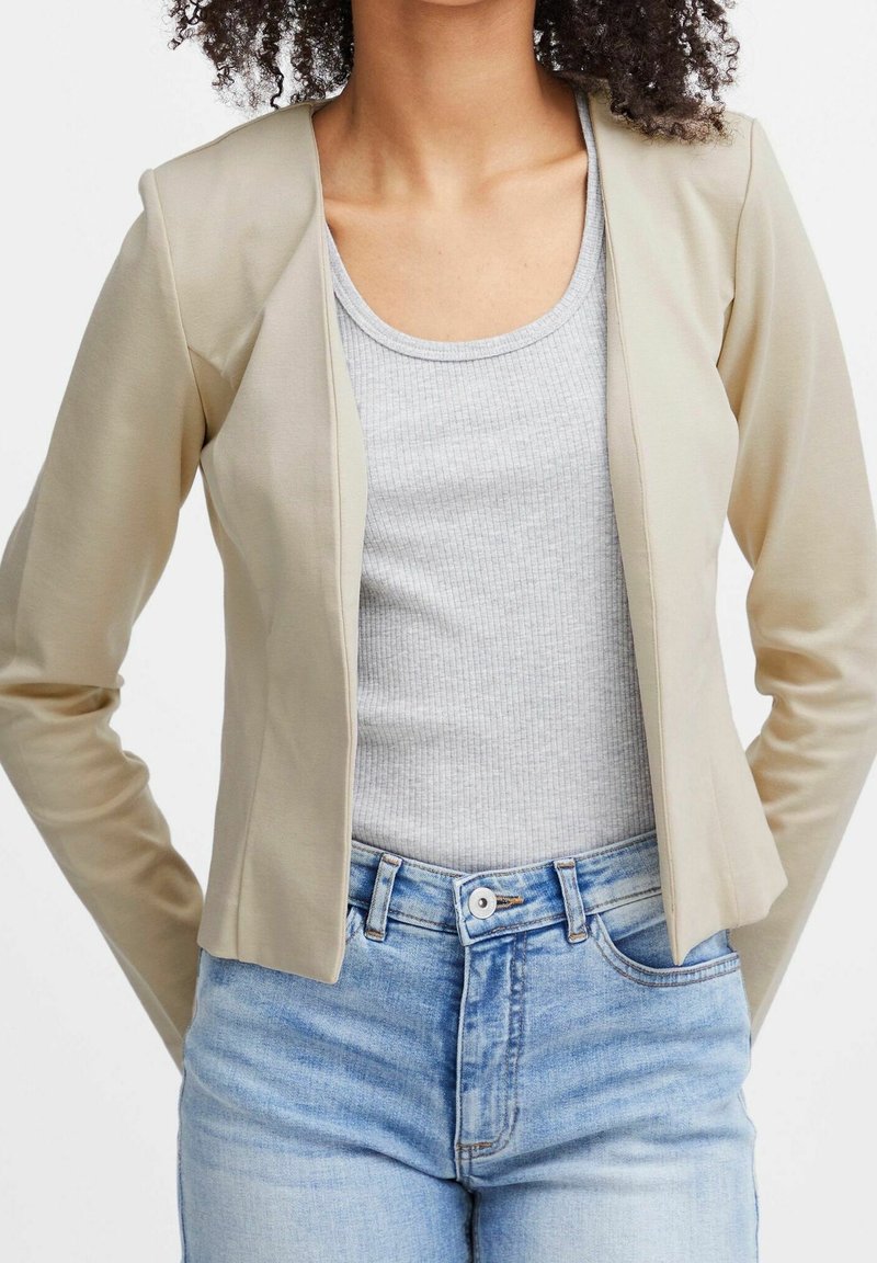 Femme portant un blazer ouvert beige sur un débardeur côtelé gris clair et un jean taille haute bleu clair, debout avec les mains derrière le dos.