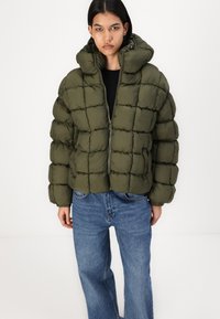 Olivengrønn pufferjakke med en boksformet silhuett, kviltet design, høy krage og glidelås foran, kombinert med blå denimjeans.