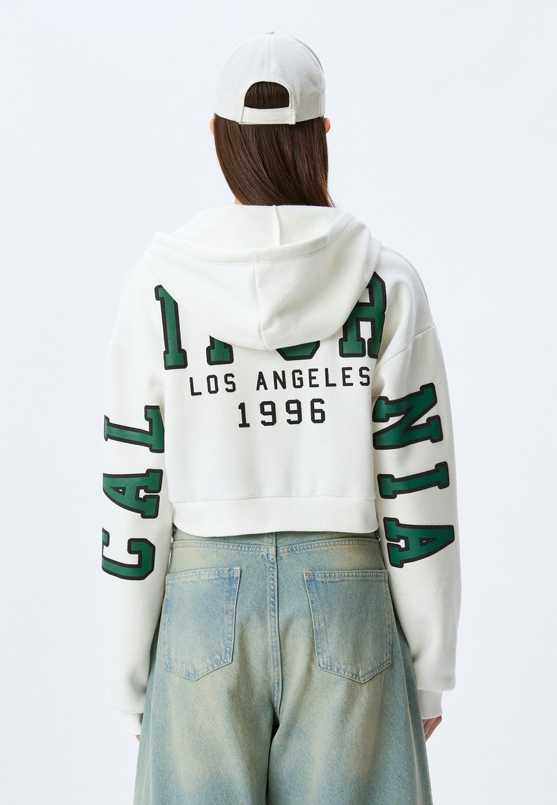 Haut à manches longues blanc avec logo vert : "CALIFORNIA," "LOS ANGELES," et "1996." Porté avec un jean évasé bleu clair.