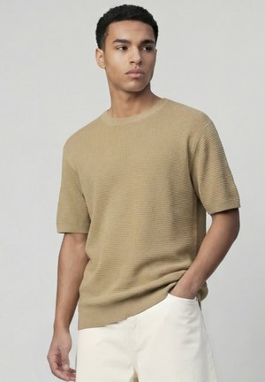 Jeune homme portant un pull beige texturé à manches courtes et un pantalon blanc, debout avec une main dans la poche, regardant sur le côté.