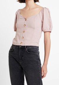 Blusa in lino rosa chiaro con maniche a palloncino, scollatura quadrata e sei bottoni di legno sul davanti. Abbinata a jeans neri a vita alta.