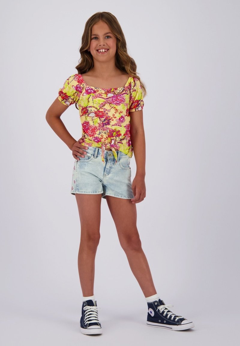 Helles gelbes Kurzarmshirt mit pinken und roten Mustern; kombiniert mit hellblauen Denim-Shorts und navyfarbenen High-Top-Sneakern.
