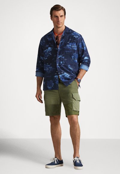 Άνδρας που φοράει μπλε πουκάμισο με μοτίβο και κουμπιά, ριγέ κόκκινη μπλούζα, πράσινα σορτς cargo, μπλε αθλητικά παπούτσια και ρολόι, στέκεται με το χέρι στην τσέπη.