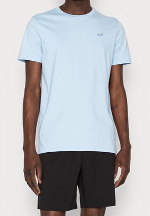 Hellblaue Baumwoll-T-Shirt mit kurzen Ärmeln, Rundhalsausschnitt und einem kleinen Logo in der oberen linken Ecke. Getragen mit schwarzen Shorts.