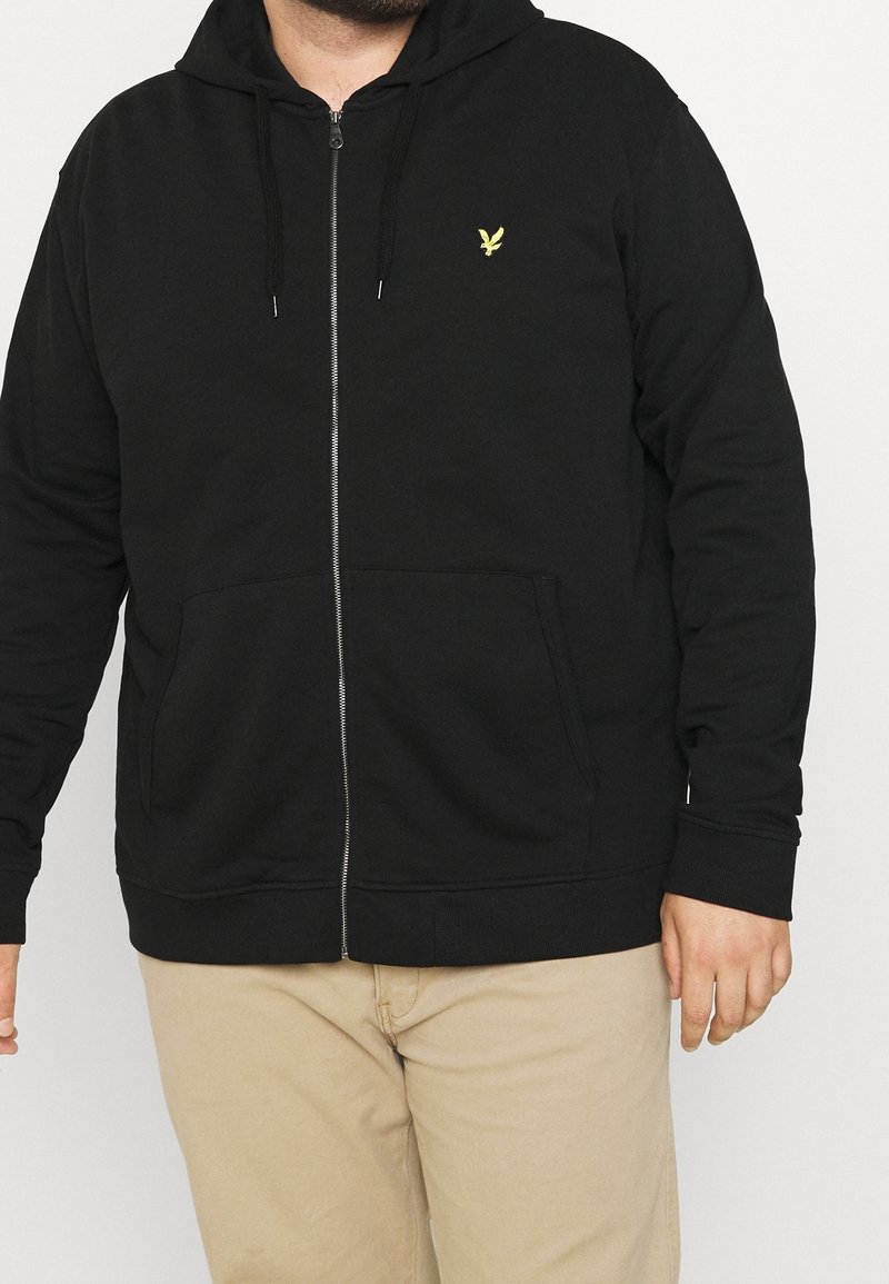 Homme portant un sweat à capuche noir zippé avec un petit logo d'oiseau jaune sur la poitrine et un pantalon beige, debout devant un fond uni.