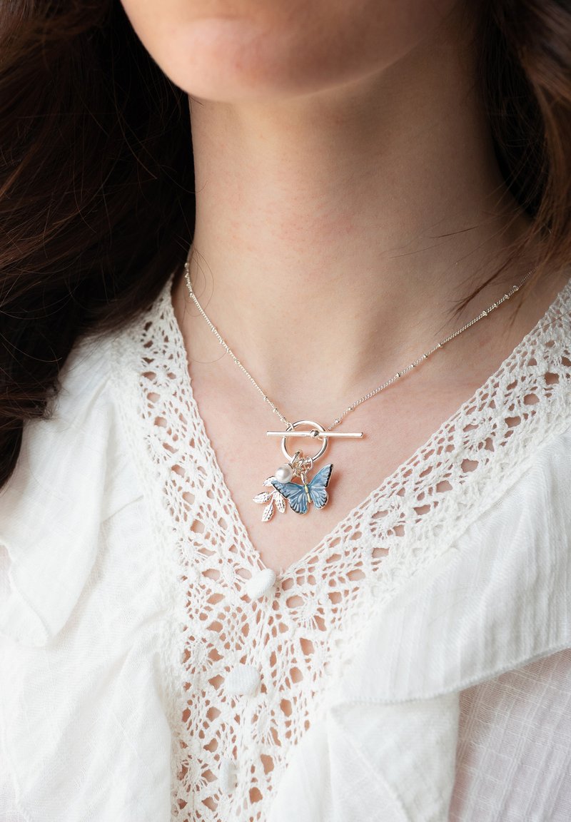 Collier en chaîne argentée avec fermoir à bascule orné de breloques papillon bleu, perle et feuille, porté sur un chemisier blanc à bordure en dentelle.