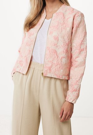Femme portant une veste zippée texturée rose clair sur une chemise blanche et un pantalon beige à jambes larges avec une taille élastique.