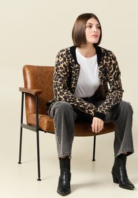 Cardigan con stampa leopardata e bordi neri, indossato sopra una t-shirt bianca. Jeans grigi e stivaletti neri. Sedia in pelle marrone sullo sfondo.