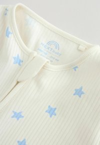 Kremasti rebrasti dječji bodi s plavim uzorcima zvjezdica i malom tkanom etiketskom trakom blizu zaobljenog zatvarača na vratu.