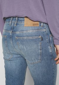 Jeans de mezclilla en azul claro con acabado desgastado. Presenta una parche de cuero marrón con la etiqueta "DRYKORN" en la cinturilla y dos bolsillos traseros.