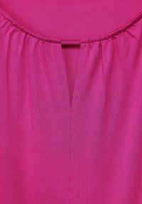 Magenta plagg med en halsringning som har en samlad elastisk kant och en liten V-formad insnitt centrerad under kragen.