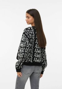 Donna con lunghi capelli castani che indossa un maglione a righe bianco e nero con la scritta "LOVE" e jeans grigi, in piedi davanti a uno sfondo bianco.