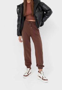 Pantaloni della tuta marroni con polsini elastici, abbinati a un top corto marrone e a un giubbotto invernale nero. Sneakers bianche e marroni completano l'outfit.