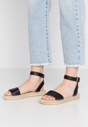 Pieds portant des sandales noires à bride cheville avec des semelles tissées, associées à un jean bleu clair à ourlet effiloché, debout sur un sol blanc.