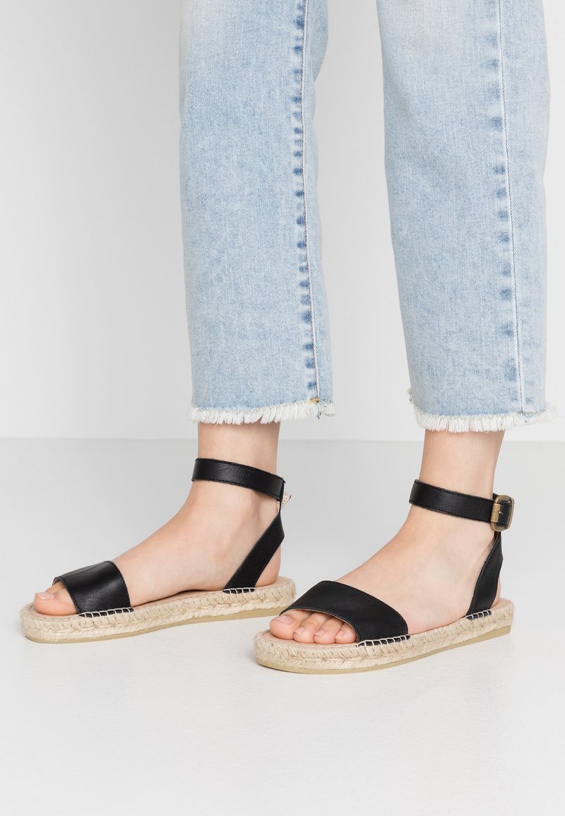 Pieds portant des sandales noires à bride cheville avec des semelles tissées, associées à un jean bleu clair à ourlet effiloché, debout sur un sol blanc.