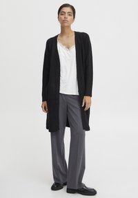 Schwarzer, langer Cardigan aus weichem, strukturiertem Stoff, mit offenem Frontbereich, über einem weißen Spitzen-Top und grauen, weiten Hosen getragen. Schwarze Schuhe.