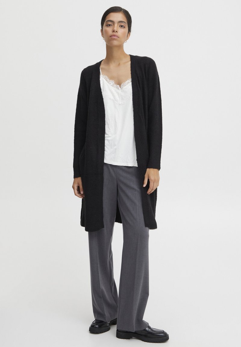 Schwarzer, langer Cardigan aus weichem, strukturiertem Stoff, mit offenem Frontbereich, über einem weißen Spitzen-Top und grauen, weiten Hosen getragen. Schwarze Schuhe.