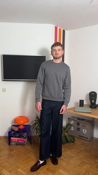 Γκρι φούτερ με στρογγυλή λαιμόκοψη, σκουρόχρωμο τζιν με φαρδιά παντελόνια και καφέ loafers με λευκές κάλτσες. Το δωμάτιο διαθέτει τηλεόραση, πορτοκαλί φωτιστικό και ξύλινο πάτωμα.