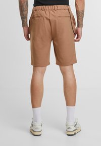 Smeđi šorts s elastičnom trakom u struku, bočnim džepovima i detaljem od lanca. Model nosi bijele čarape i New Balance tenisice. Glatka tekstura materijala.