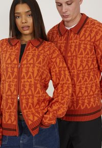 Pulls zippés en tricot de couleur orange vif, avec un motif géométrique, des accents noirs au col et aux poignets, et une coupe décontractée.