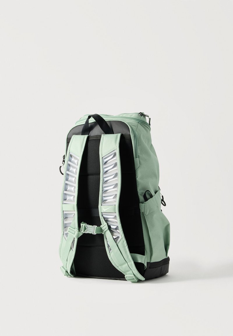 Mochila verde claro con panel trasero acolchado negro, correas de hombro ajustables con detalles plateados reflectantes y bolsillos laterales.