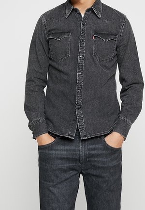 Mężczyzna ubrany w wyblakłą czarną jeansową koszulę z dwoma kieszeniami na piersi oraz pasujące czarne jeansy, ręce w kieszeniach, stoi na jednolitym tle.