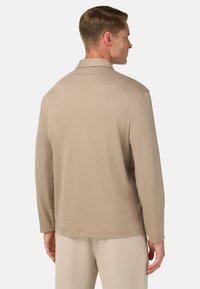 Camicia beige a maniche lunghe con una texture morbida, vestibilità rilassata e colletto classico, vista da dietro su uno sfondo neutro.