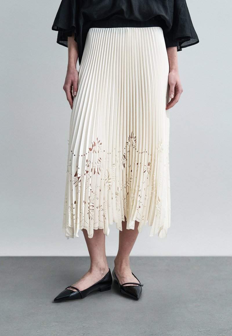 Κρεμ φούστα με πιέτες και λουλουδάτες κοπές στο τελείωμα, συνδυασμένη με μαύρες μυτερές μπαλαρίνες. Η φούστα έχει υφασμάτινο, ροής σχεδιασμό.