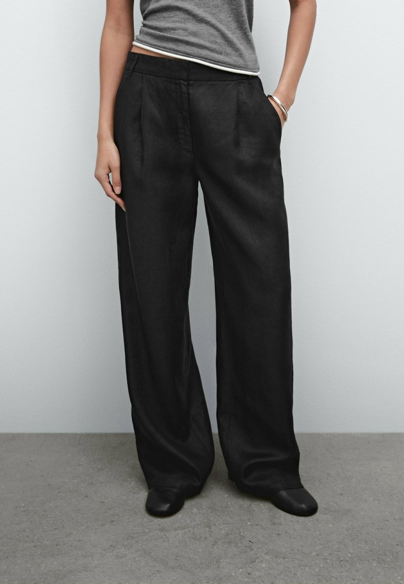 Personne portant un pantalon large noir ample, un haut gris sans manches et des chaussures noires, debout avec une main dans la poche sur un fond uni.
