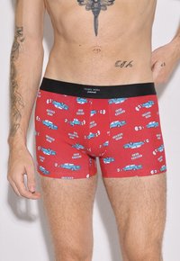 Hombre con calzoncillos rojos con estampado de un coche azul y "Las Vegas", cinturón negro con la etiqueta "Ysabel Mora", tatuajes visibles en brazos y torso.