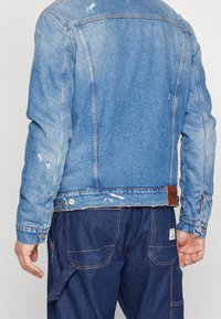 Ljusblå denimjacka med slitna detaljer, knäppta ärmslut och en läderetikett vid fållen. Bärs med mörkblå cargo-byxor.