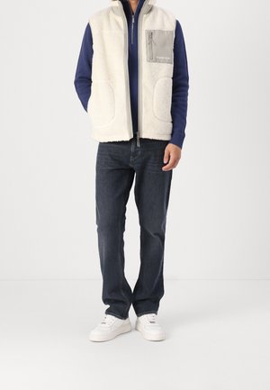 Chaleco de forro polar en color crema con un bolsillo de pecho con cremallera gris, llevado sobre un suéter azul marino, combinado con jeans oscuros y zapatillas blancas.
