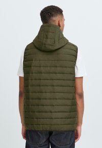 Olivegrønn polstret vest med hette, med horisontal quilt og lett tekstur, designet uten ermer for bevegelsesfrihet.