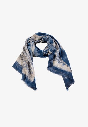 Blåt mønstret tørklæde med vævet design og frynsede kanter, elegant draperet. Model iført let top og teal bukser, barfodet, mod en neutral baggrund.