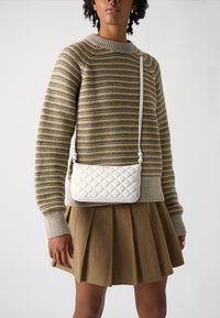 Borsa a tracolla bianca trapuntata con finitura liscia, dotata di una lunga tracolla e di accessori in metallo argentato, indossata con un maglione a righe e una gonna plissettata.