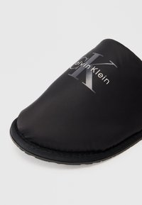 Čierne nazúvacie papuče s gumovou podrážkou, polstrovanou vrchnou časťou a bielym a sivým logom Calvin Klein vytlačeným na vrchu.