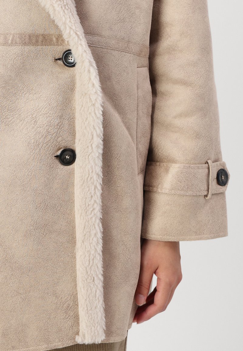 Manteau en suède synthétique beige, doté d'un doux col et d'une doublure en fausse fourrure crème, de grands boutons noirs et de manches retroussées avec des boutons assortis.