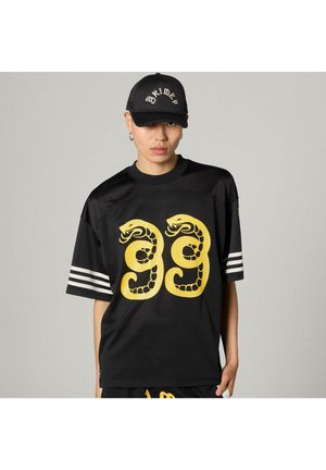 Giovane con cappellino nero e maglia nera con il numero 99 disegnato a forma di serpente giallo e strisce bianche sulle maniche, in piedi davanti a uno sfondo grigio.